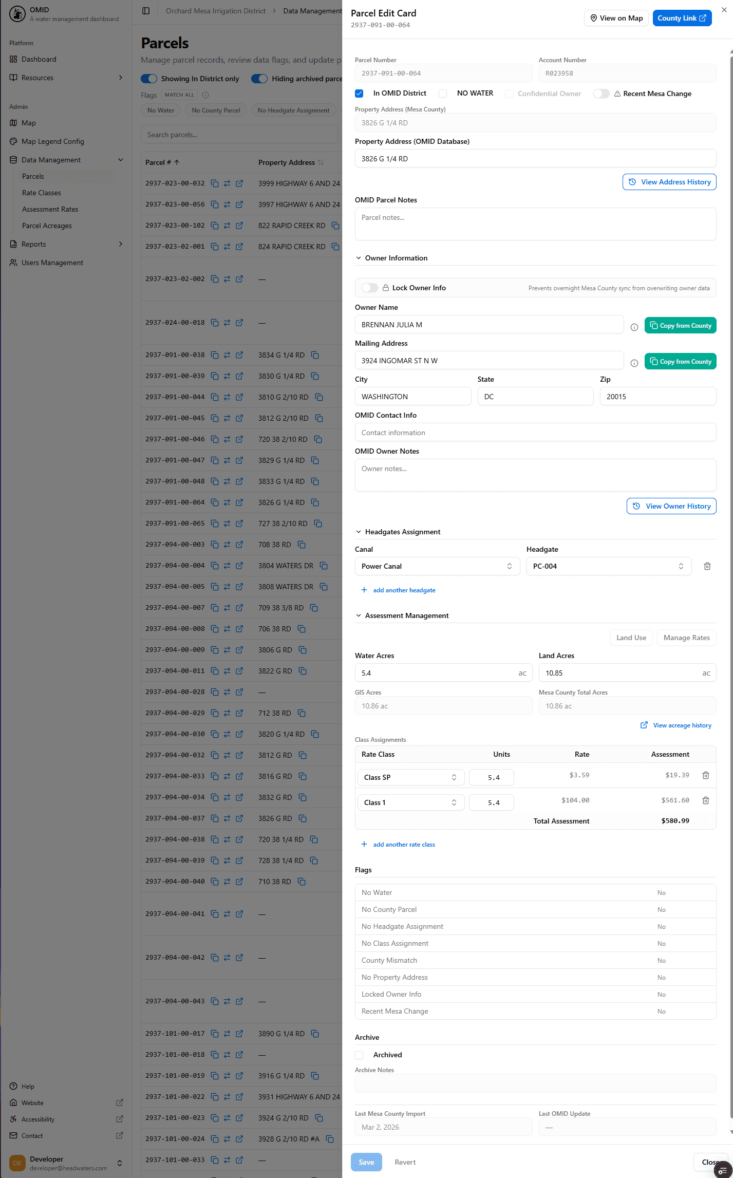 Rich custom Parcel data management interface (OMID). A left nav organizes the admin app (Dashboard, Map, Map Layers Config, and Data Management sub-modules: Parcels, Rate Classes, Assessment Rates, Parcel Acreage, Reports, User Management). The main list is a searchable, filterable parcel grid (parcel #, property address) with per-row actions and toggles like 'Showing in District only' and 'Hiding archived parcels'. A detailed Parcel Edit Card on the right edits a single record across sections: Property Address (with Mesa County database sync), Owner Information (lockable owner info, mailing address, OMID contact/notes, copy-from-county, view owner history), Headgate Assignment (canal + headgate), Assessment Management (water/land acres plus a rate-class table with units/rate/assessment and total), boolean Flags, Archive, and county/OMID import timestamps, with Save/Revert. Demonstrates deep, domain-specific CRUD with external county data integration, audit history and rich validation.